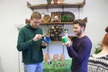 A couple of male students painting pots in front of a supply table